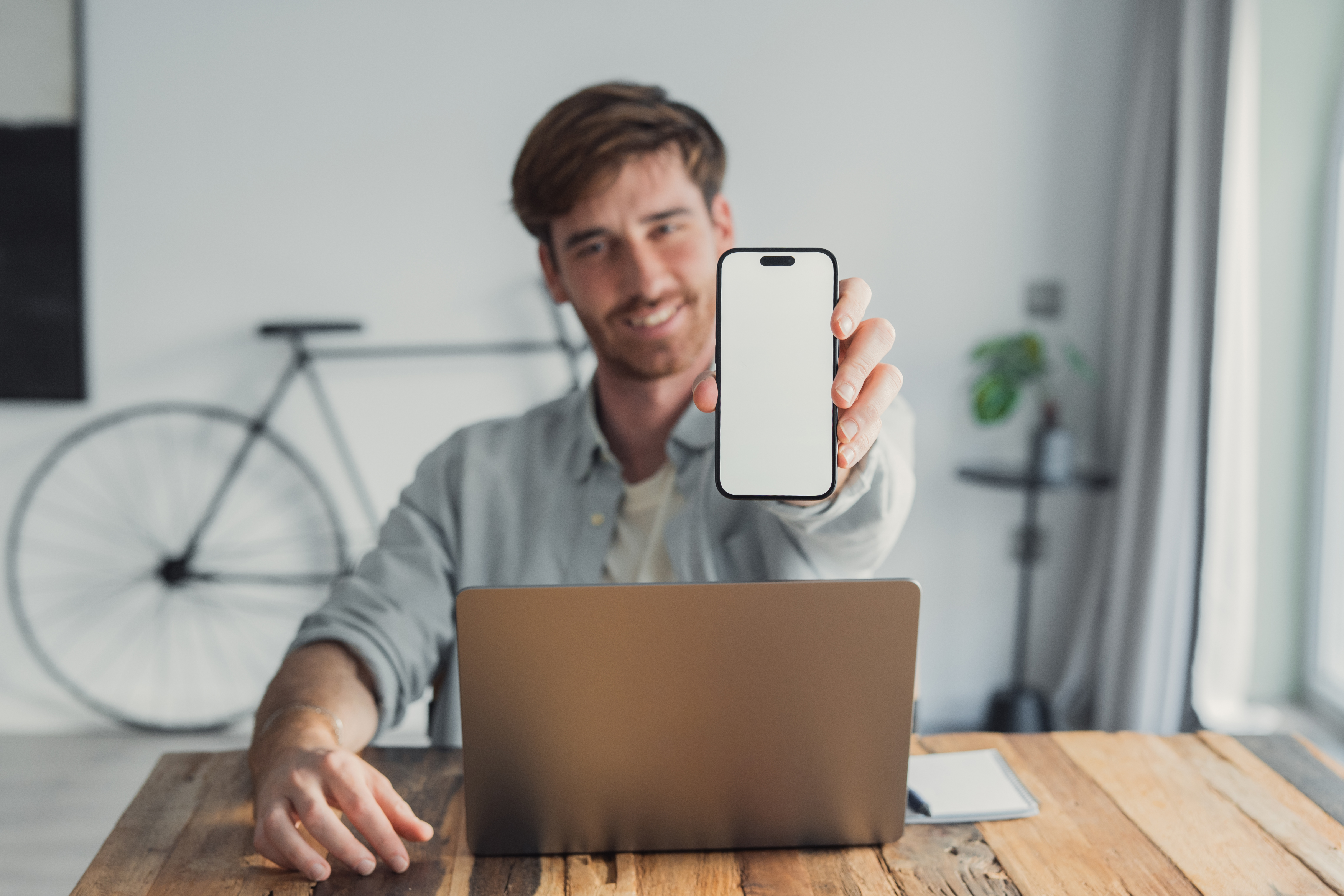 Young man smiling at camera while working from home, holding a smartphone with a blank screen for app or website mockup, casual home office scene ready for product placement
