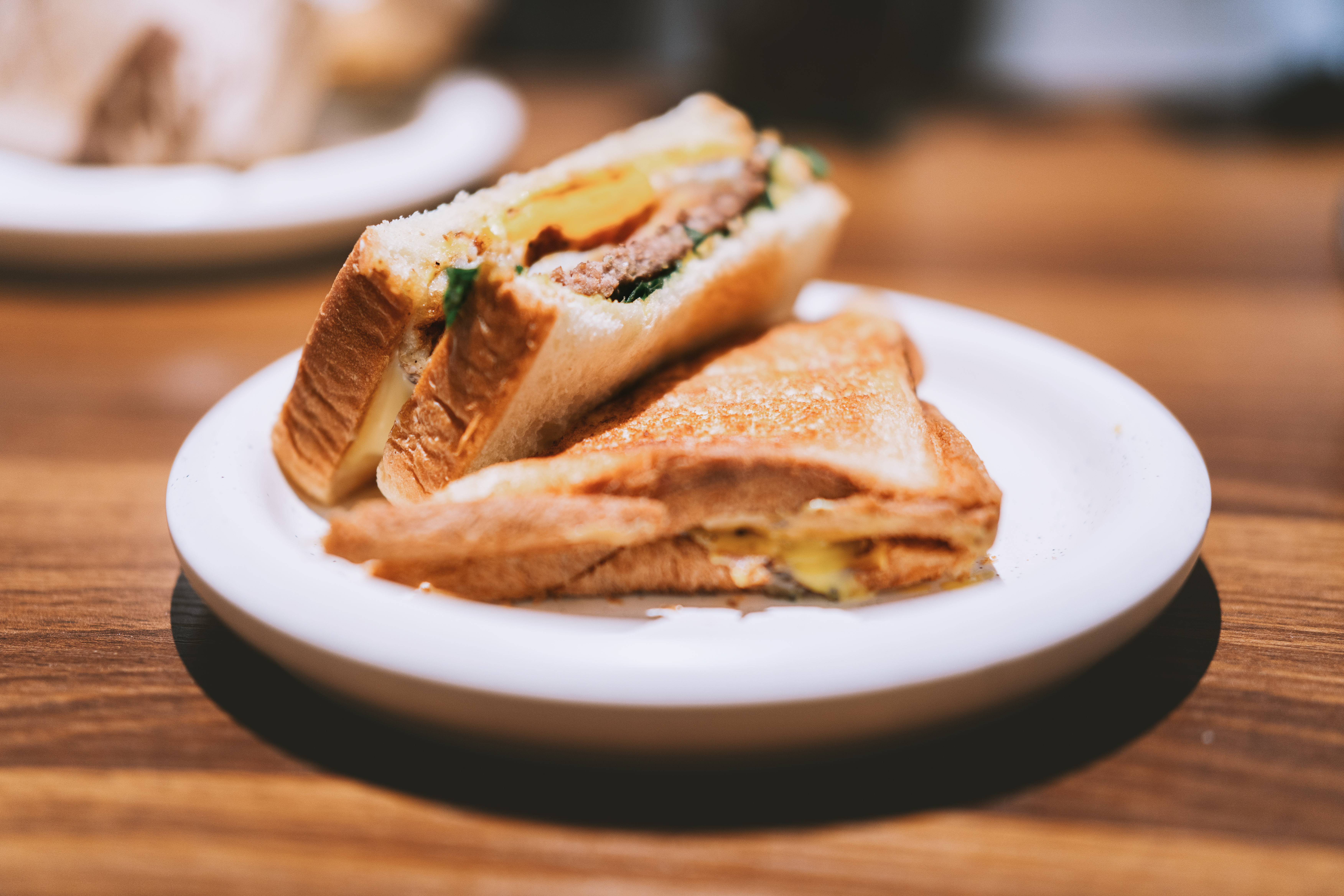 Grilled cheese and egg sandwich with sausage, served on white plate atop wooden table in cozy café
